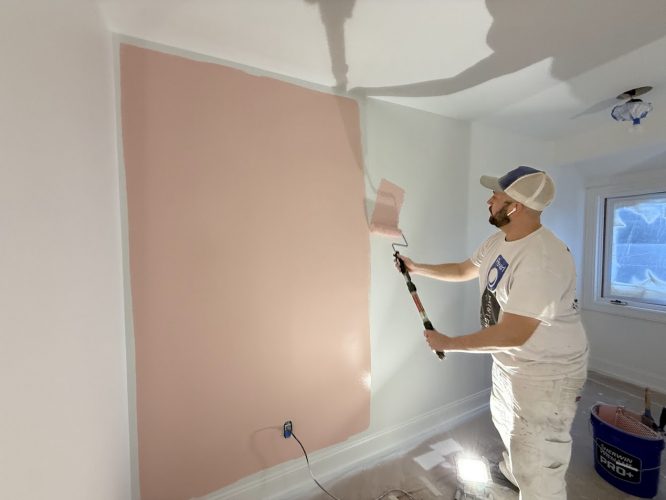 Painter in white clothes paints a white wall light pink with a roller; unfurnished, bright room. Pearl Painters, Portland.