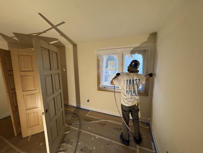 Painter in cap and white shirt spray-paints window frame in taped, mostly empty room. Pearl Painters at work in Portland.