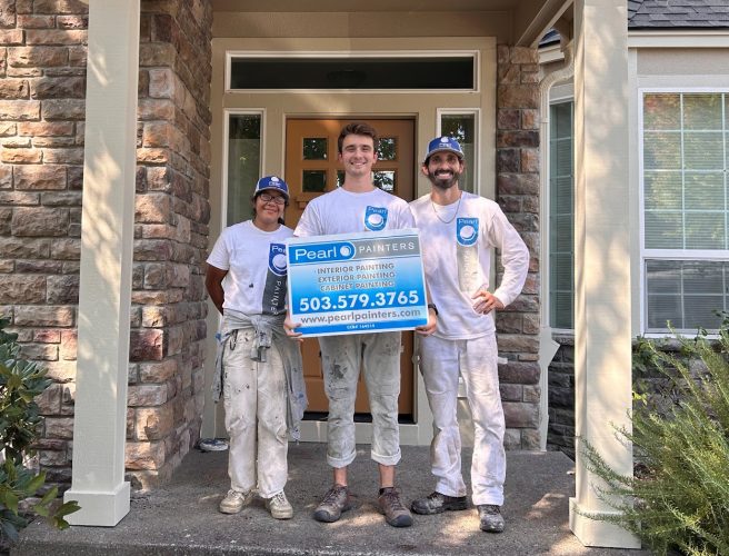 Three painters in white outfits stand by a house in Portland, holding a Pearl Painters sign with contact details.