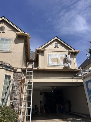 A painter from Pearl Painters paints a house exterior in Portland, with windows and trim protected by plastic and tape.