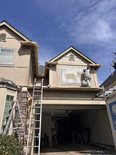 A painter from Pearl Painters paints a house exterior in Portland, with windows and trim protected by plastic and tape.