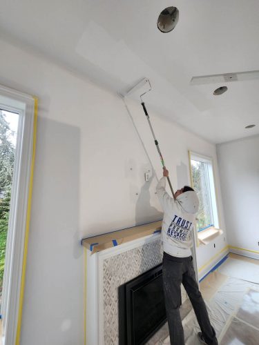 A painter from Pearl Painters in Portland skillfully uses an extended roller to paint a living room ceiling white, with the floor protected.