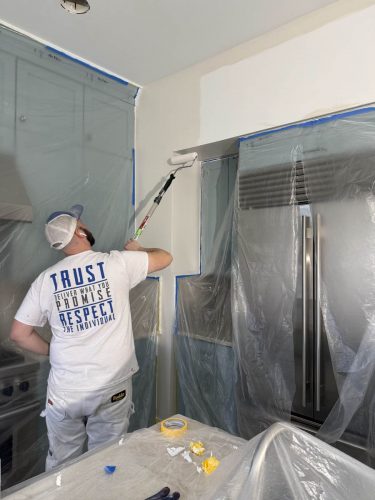 A painter in Portland uses a roller to paint the kitchen wall, protecting the interior with plastic sheets.