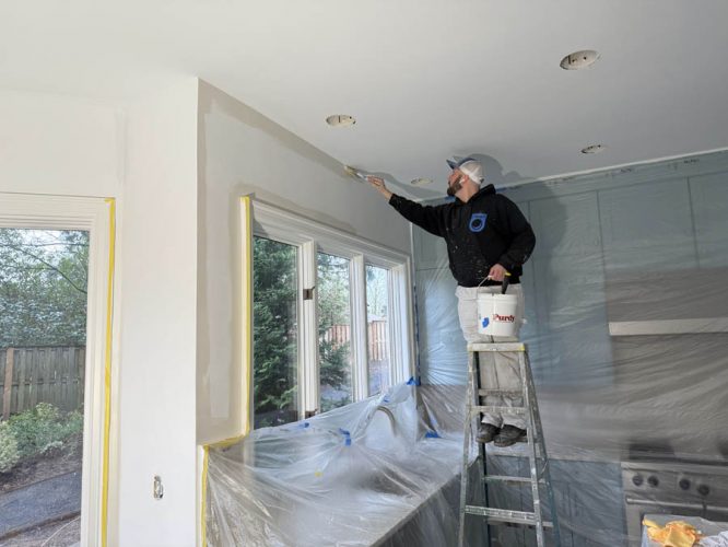 A painter on a ladder painting the upper edge of a beige kitchen wall. Interior surfaces and appliances are covered with protective plastic sheets to keep everything clean.