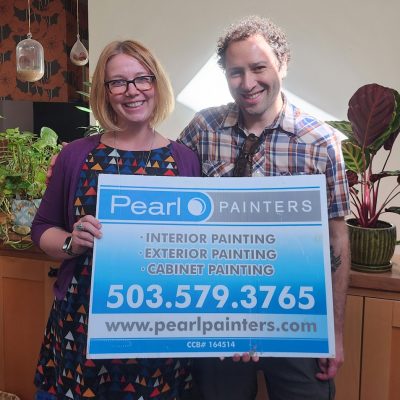 Two people indoors hold a Pearl Painters sign for painting services in Portland; plants are in the background.