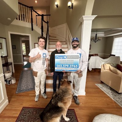 Three men in a home entryway, two holding a Pearl Painters painting sign. German Shepherd, staircase, and covered furniture visible.