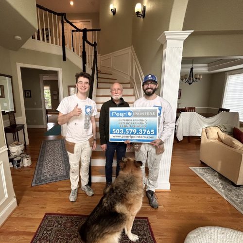 Three men in a home entryway, two holding a Pearl Painters painting sign. German Shepherd, staircase, and covered furniture visible.
