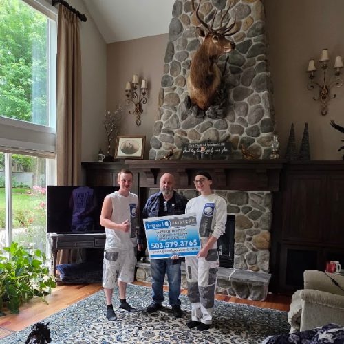 Three people hold a Pearl Painters sign in a living room with a stone fireplace and mounted deer head, likely related to painting services in Portland.