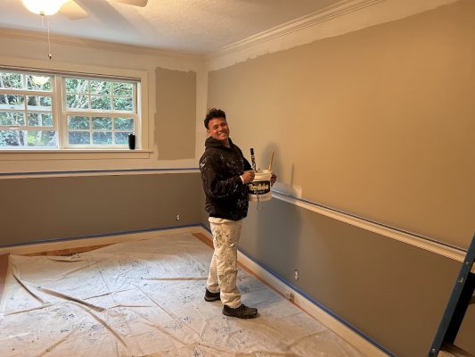 A painter with supplies stands in a room with drop cloths, walls partially primed, ready for painting by Pearl Painters in Portland.
