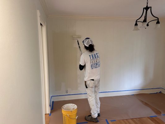 Painter uses roller to paint cream wall; floor is covered for protection, yellow bucket nearby. Pearl Painters in Portland.