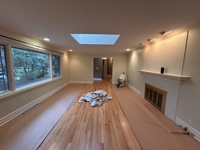 Painter preps living room with wood floors, covered surfaces, skylight, big windows, and fireplace for Pearl Painters in Portland.