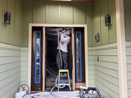 Painter on step ladder uses power tool on plastic-covered door frame, with painting supplies and taped windows. Pearl Painters, Portland.