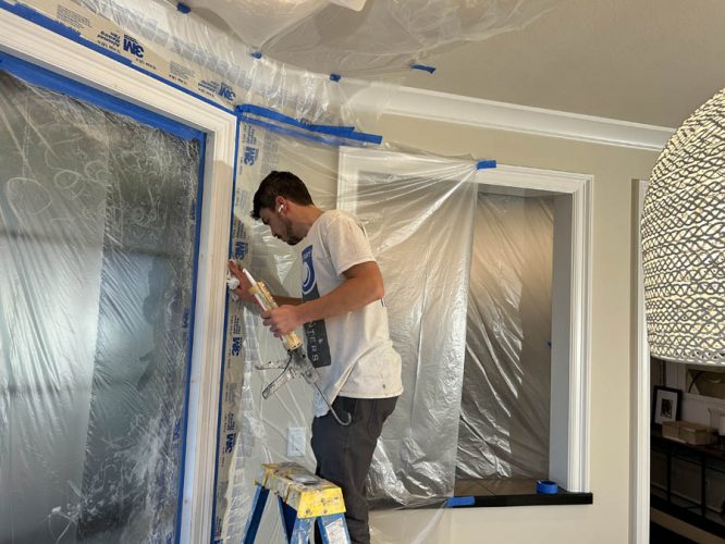A painter for Pearl Painters is caulking a window frame in West Linn, with walls covered by plastic sheets.
