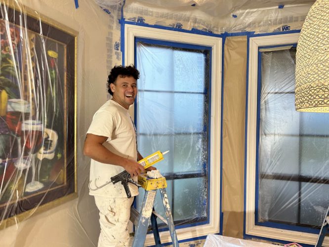 A painter in white work clothes stands on a ladder with a caulk gun and container. The room is meticulously prepared with protective plastic and taped windows for painting, showcasing Pearl Painters' high precision in West Linn.