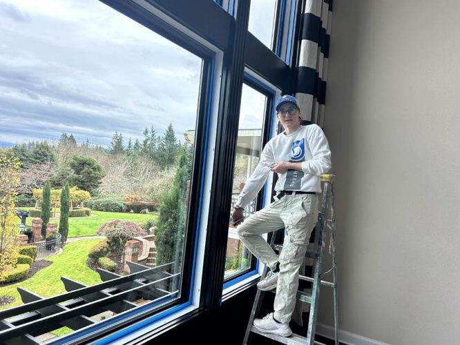 A painter from Pearl Painters works on the interior near blue-framed windows overlooking a garden in West Linn.