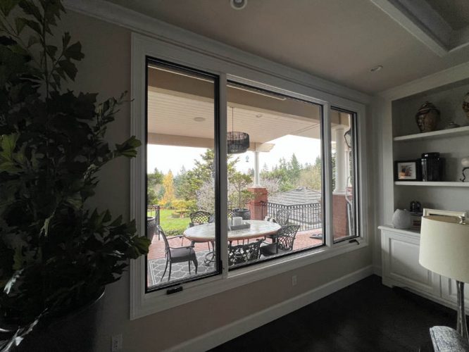 Through a large window in West Linn, the patio shows an inviting outdoor table and chairs surrounded by greenery