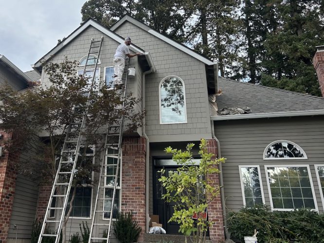 Painter on a ladder painting the upper wall of a two-story brick and gray house, trees around, Pearl Painters, Portland.