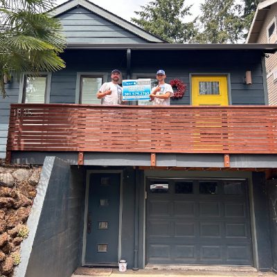 Two people stand on the balcony of a gray house with a yellow door in Portland, holding a Pearl Painters sign with contact info.