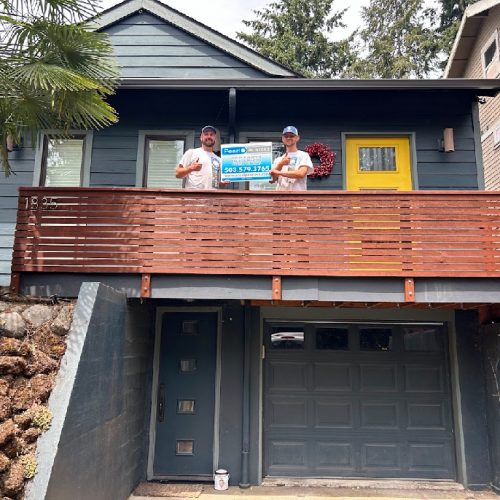 Two people stand on the balcony of a gray house with a yellow door in Portland, holding a Pearl Painters sign with contact info.