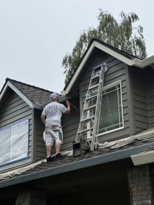 A painter paints house trim on a roof near a ladder; windows are covered. Pearl Painters at work in Portland.