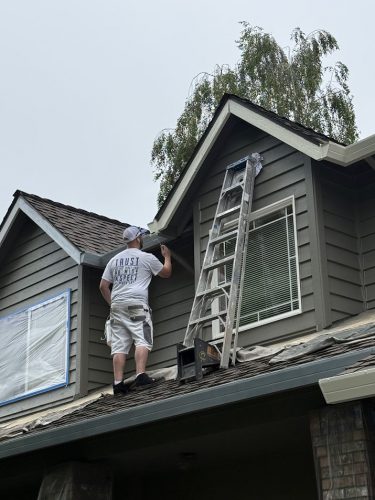 A painter paints house trim on a roof near a ladder; windows are covered. Pearl Painters at work in Portland.