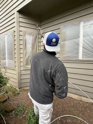 A painter in paint-stained clothes sprays paint on a house exterior with windows covered in plastic. Pearl Painters, Portland.