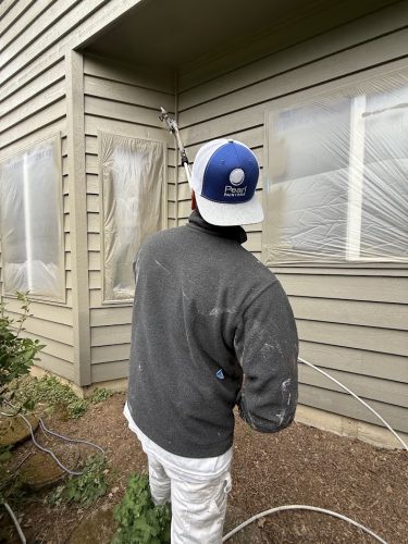 A painter in paint-stained clothes sprays paint on a house exterior with windows covered in plastic. Pearl Painters, Portland.