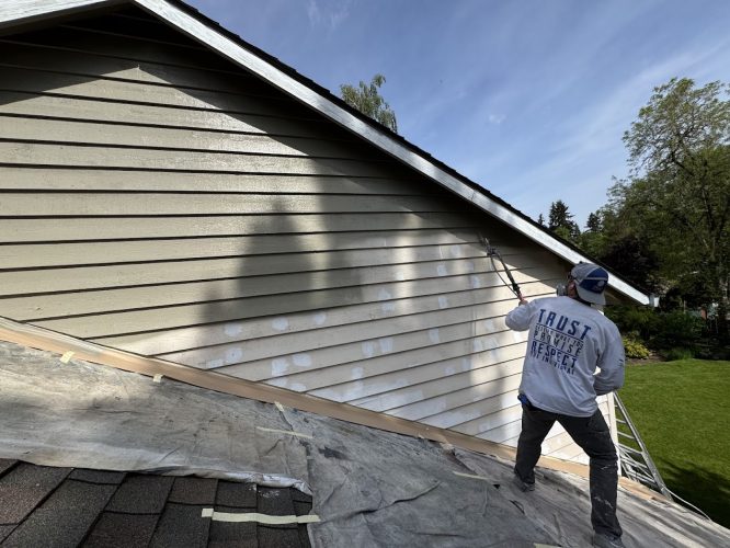 A painter on a sloped roof uses a roller to paint a house beige; prep patches and drop cloths are visible. Pearl Painters, Portland.