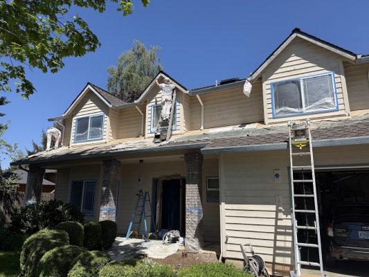 Painters from Pearl Painters paint a beige two-story house exterior in Portland using ladders and scaffolding on a sunny day.