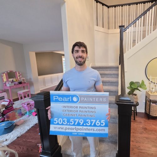 Man indoors in Portland holding Pearl Painters sign for painting; contact info, staircase, and kids' play area visible behind him.