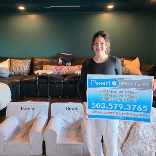Woman with Pearl Painters sign stands in Portland living room by dog beds labeled Bardot and Brody, in front of sectional sofa.