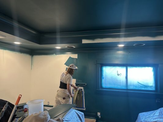 A painter from Pearl Painters in Portland rolls teal paint on a wall; furniture is covered and windows are taped for protection.