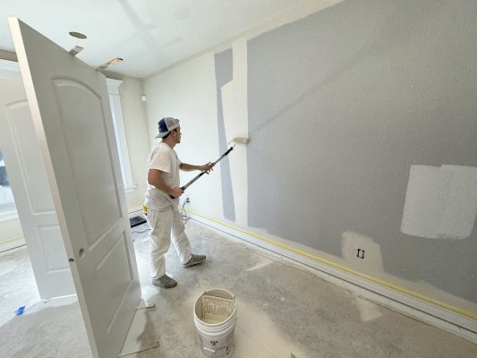 A painter in white work clothes paints a wall light gray with a roller; room under renovation by Pearl Painters in Portland.
