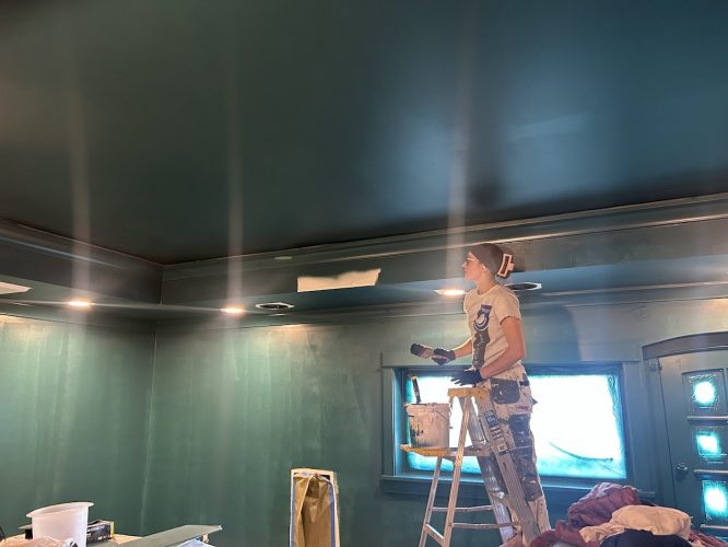 A painter on a ladder paints a teal wall and ceiling, with supplies and lights around. Pearl Painters, Portland.