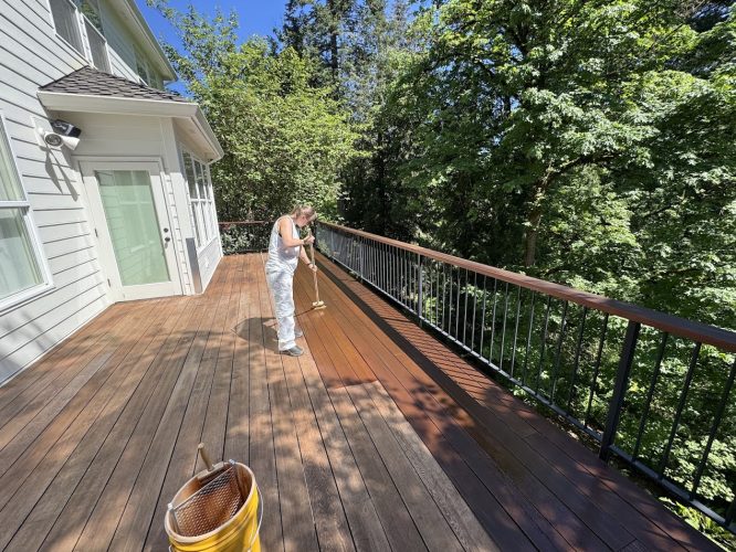 A painter stains a wooden deck with a brush outside a house, trees in the background, on a sunny day. Pearl Painters, Portland.