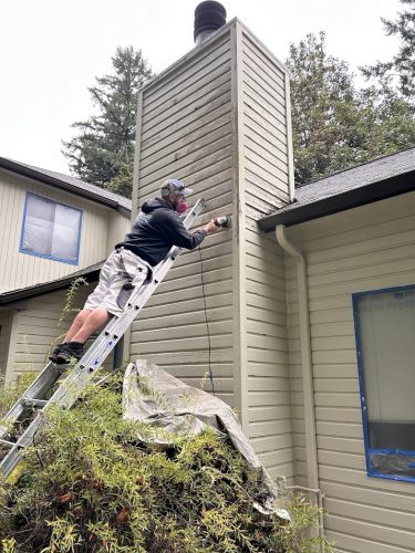 Painter in safety gear stands on a ladder using a power tool on a house chimney. Pearl Painters, Portland.