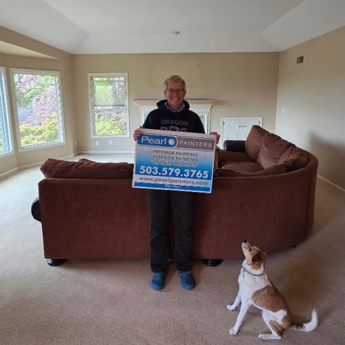 Person in a living room in Portland holding a Pearl Painters sign, with a brown sectional and dog nearby; related to painting.