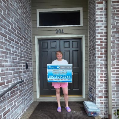Person holding Pearl Painters painting sign by door 204, brick walls and welcome mat. Scene appears to be in Portland.