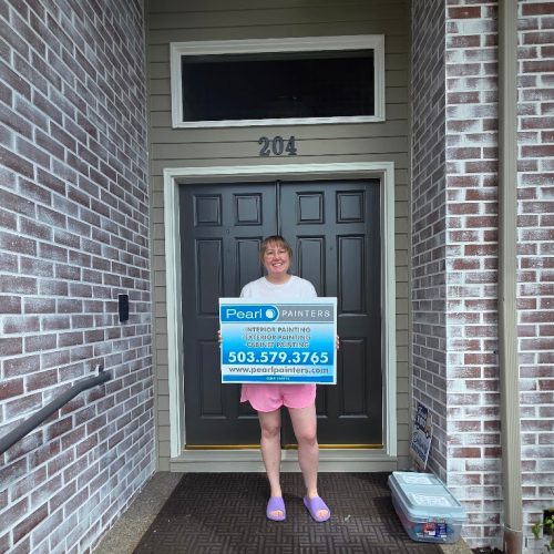 Person holding Pearl Painters painting sign by door 204, brick walls and welcome mat. Scene appears to be in Portland.