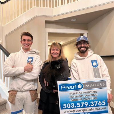Two painters and a woman with a dog stand indoors by stairs, holding a Pearl Painters sign. Portland painting service.