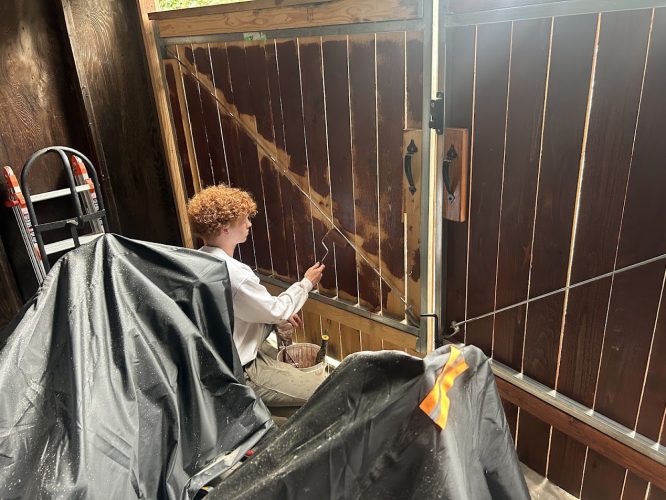Painter kneeling and painting a wooden gate with a brush; ladder and covered items nearby, likely for Pearl Painters in Portland.