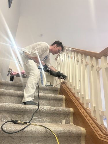 A painter sands a wooden stair railing in Portland; plastic covers carpeted stairs. Pearl Painters is on the job.