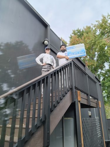 Two people stand atop outdoor stairs by a black building in Portland, holding a blue and white Pearl Painters sign; trees behind.