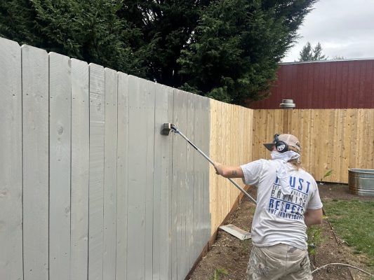 A painter is painting a wooden fence gray with a roller brush outdoors in Portland; some boards on the right are unpainted.