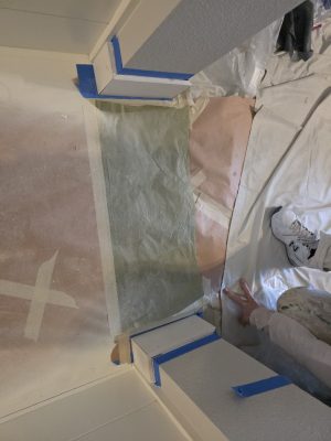 Painter taping brown and green paper to walls and floor in a corner, prepping for painting; white sneakers, drop cloth visible.
