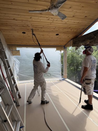 Painters from Pearl Painters in Portland spray-paint a wooden porch ceiling with plastic sheeting protecting surfaces.