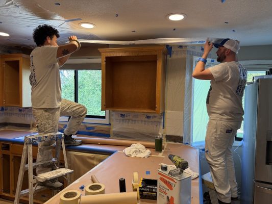 Two painters in white tape plastic sheeting to a kitchen ceiling and cabinets, prepping for painting. Pearl Painters, Portland.