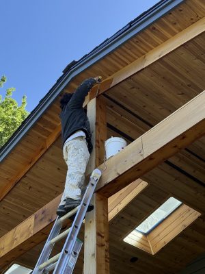 Painter on a ladder painting the exterior wooden house structure under clear blue sky, representing Pearl Painters in Portland.