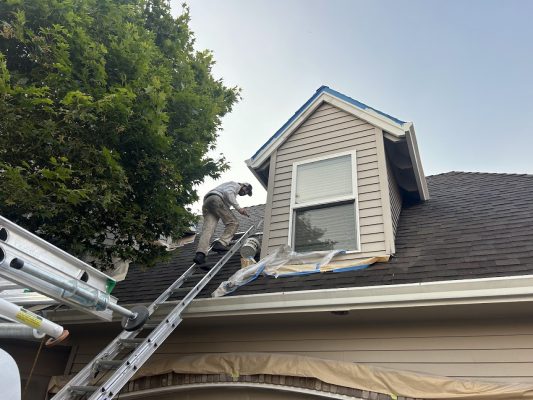 A painter on a ladder paints window trim with plastic sheeting below for protection. Pearl Painters at work in Portland.