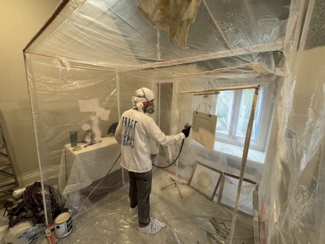 Painter in protective gear spray-paints a panel in a plastic enclosure; painting supplies on table. Pearl Painters, Portland.